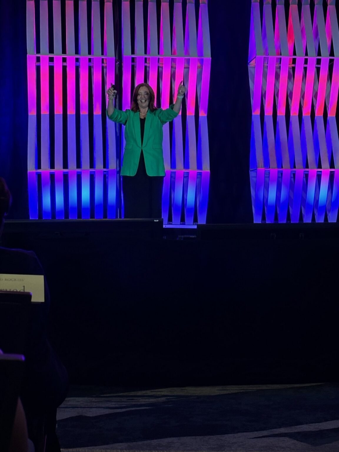 Michelle Prince keynoting to a large audience under purple and magenta stage lighting, arms raised.