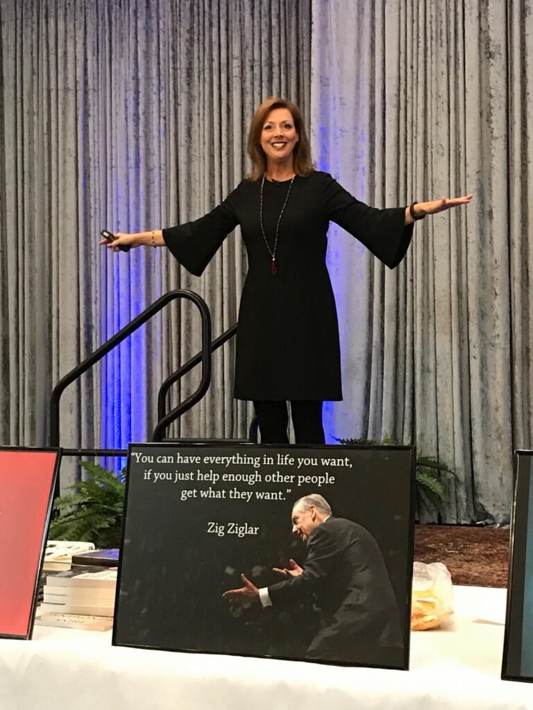 Michelle Prince on stage beside a banner of Zig Ziglar and his quote about helping other people.